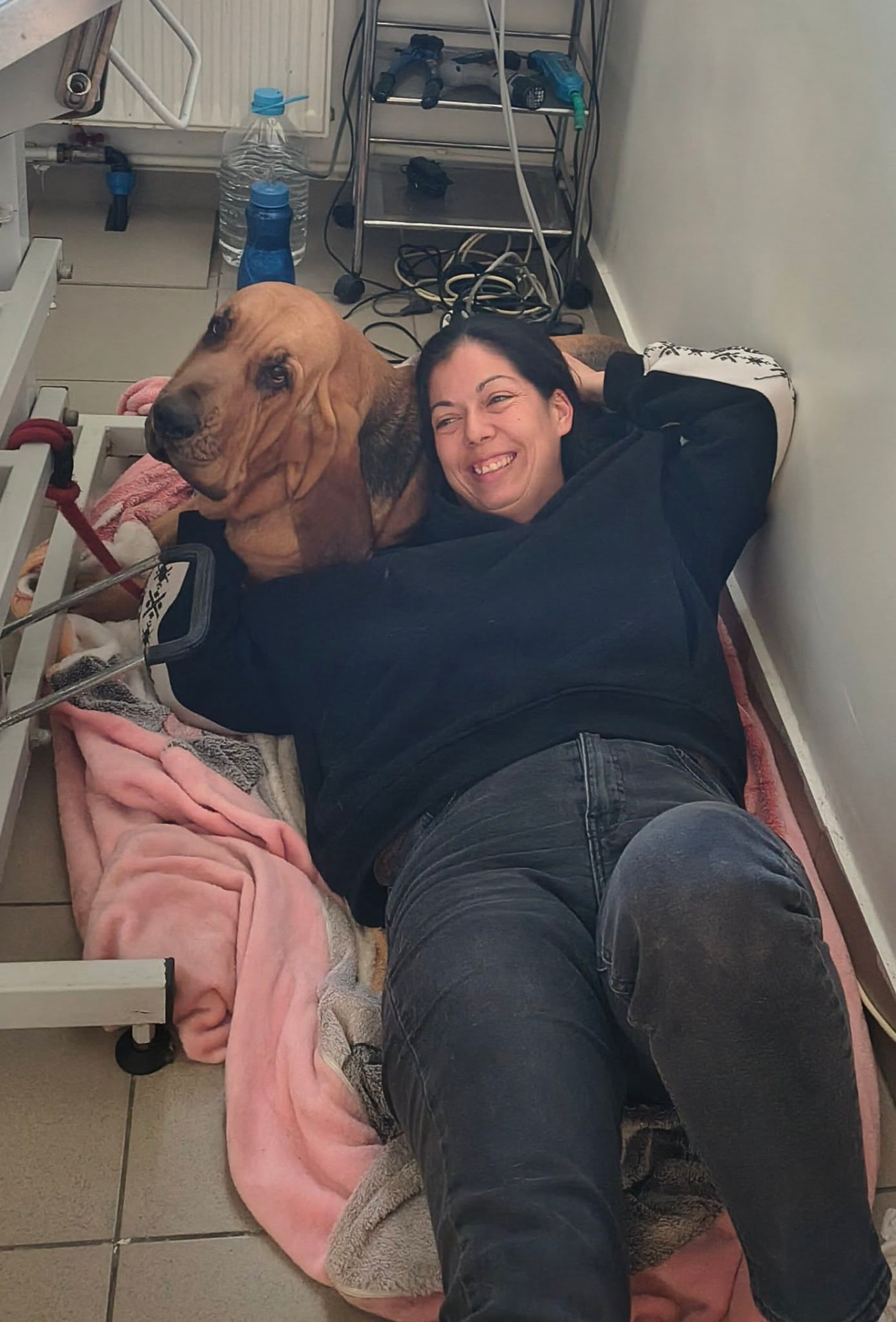 O femeie zâmbitoare stă întinsă pe o pătură lângă un câine de rasă Bloodhound, într-o încăpere interioară